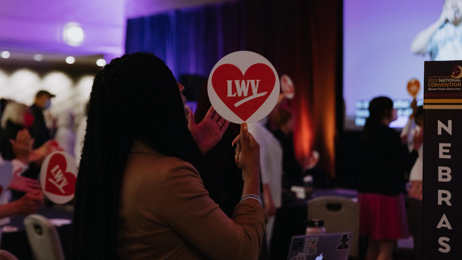 A photograph of a woman in profile at Convention 2022 in Denver, CO. The woman is in the plenary ballroom holding a LWV paddle - the paddle is white with a red heart, and the white LWV logo in the middle. She is standing up, with a NEBRASKA sign on the table next to them.